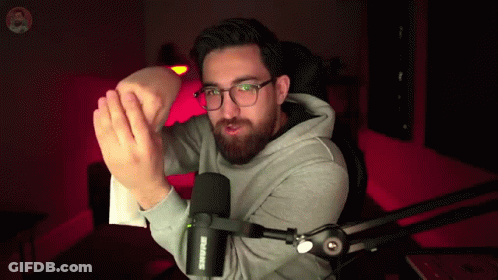 A Man Wearing Glasses And A Grey Hoodie Is Standing In Front Of A Shure Microphone GIF