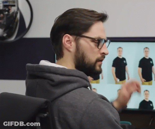 A Man Wearing Glasses And A Grey Hoodie Looks At A Monitor GIF