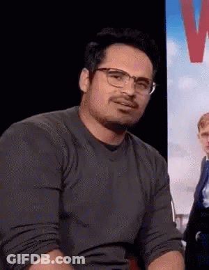 A Man Wearing Glasses And A Grey Shirt Is Sitting In Front Of A Sign . GIF