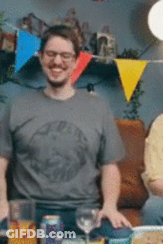 A Man Wearing Glasses And A Grey Shirt Is Standing In Front Of A Table GIF
