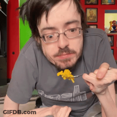 A Man Wearing Glasses And A Grey Shirt With The Letter S On It Is Eating Food GIF
