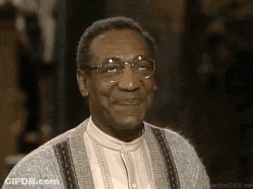 A Man Wearing Glasses And A Grey Sweater Is Smiling GIF