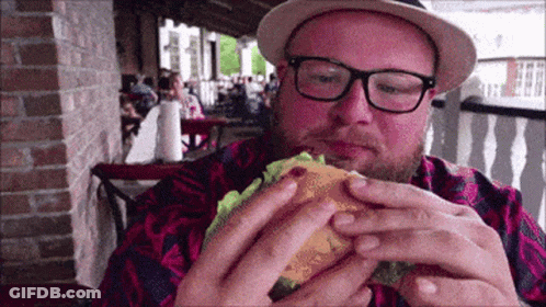 A Man Wearing Glasses And A Hat Is Eating A Burger GIF