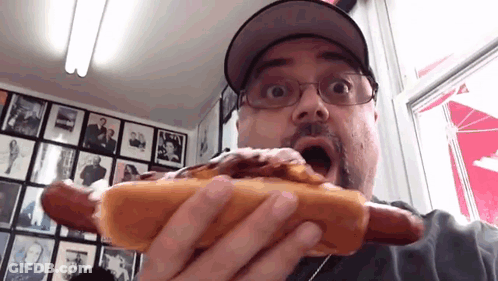 A Man Wearing Glasses And A Hat Is Eating A Hot Dog With A Surprised Look On His Face GIF