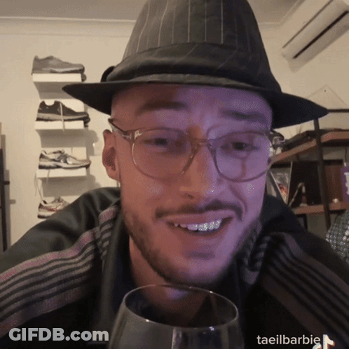 A Man Wearing Glasses And A Hat Is Holding A Glass Of Wine GIF