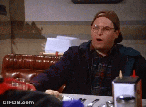 A Man Wearing Glasses And A Hat Is Sitting At A Table In A Diner With A Sandwich On It . GIF