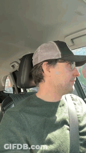 A Man Wearing Glasses And A Hat Is Sitting In A Car GIF