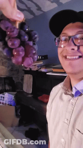 A Man Wearing Glasses And A Hat Is Smiling While Holding A Bunch Of Grapes GIF