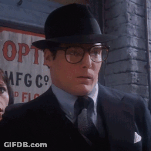 A Man Wearing Glasses And A Hat Is Standing In Front Of A Sign That Says Opti Mfg Co GIF