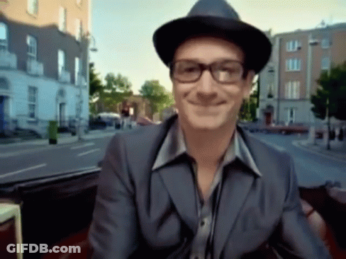 A Man Wearing Glasses And A Hat Smiles While Sitting In A Car GIF