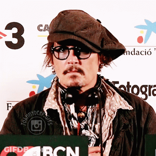 A Man Wearing Glasses And A Hat Stands In Front Of A Bcn Sign GIF