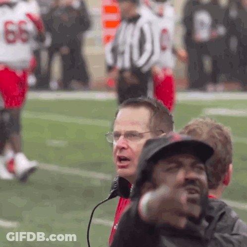 A Man Wearing Glasses And A Hat Stands On A Football Field In Front Of A Referee With The Number 66 On His Jersey GIF