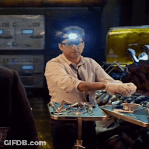 A Man Wearing Glasses And A Head Light Stands In Front Of A Table With Surgical Instruments On It GIF