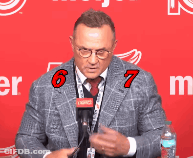 A Man Wearing Glasses And A Lanyard With The Number 6 And 7 On It Stands In Front Of A Red Background GIF