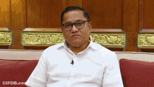 A Man Wearing Glasses And A Microphone Sits On A Red Couch GIF