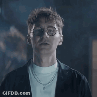 A Man Wearing Glasses And A Necklace Is Looking At The Camera In A Dark Room . GIF