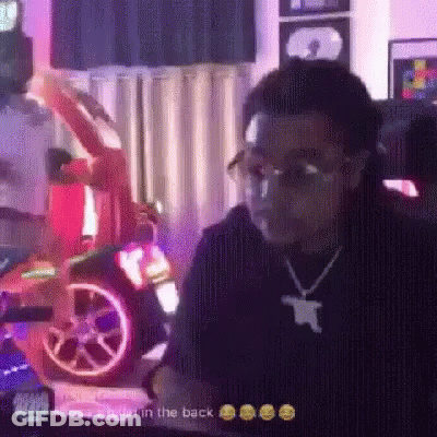A Man Wearing Glasses And A Necklace Is Sitting In Front Of A Car . GIF