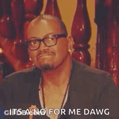 A Man Wearing Glasses And A Necklace Is Sitting In Front Of A Wooden Railing And Making A Funny Face . GIF