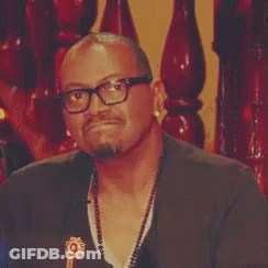 A Man Wearing Glasses And A Necklace Is Sitting In Front Of A Wooden Staircase . GIF