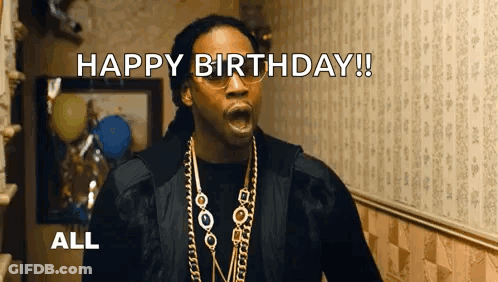 A Man Wearing Glasses And A Necklace Is Standing In A Hallway . GIF