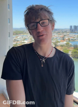 A Man Wearing Glasses And A Necklace Stands In Front Of A View Of A City GIF