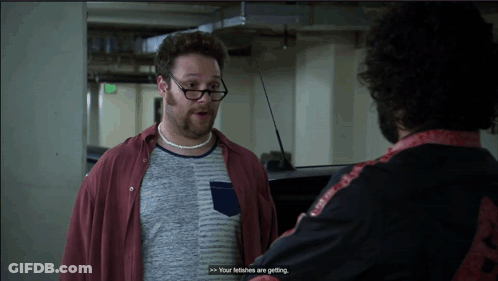 A Man Wearing Glasses And A Necklace Talks To Another Man In A Parking Garage GIF