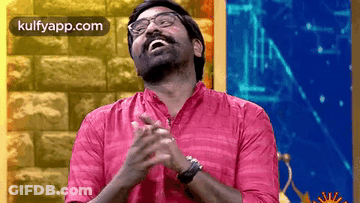 A Man Wearing Glasses And A Pink Shirt Is Clapping His Hands In Front Of A Brick Wall . GIF