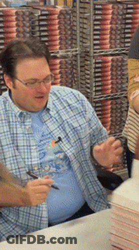 A Man Wearing Glasses And A Plaid Shirt Is Signing Books GIF