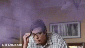 A Man Wearing Glasses And A Plaid Shirt Is Sitting In A Room With Purple Smoke Coming From His Head . GIF