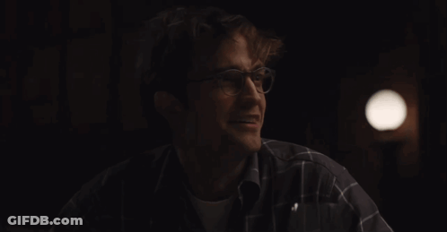 A Man Wearing Glasses And A Plaid Shirt Smiles In The Dark GIF