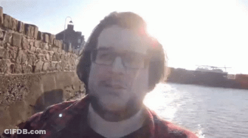 A Man Wearing Glasses And A Plaid Shirt Stands In Front Of A Body Of Water GIF
