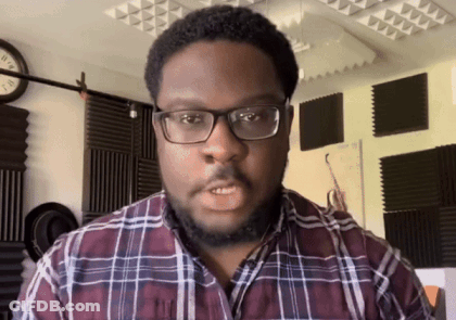 A Man Wearing Glasses And A Plaid Shirt Stands In Front Of A Wall With A Clock On It GIF