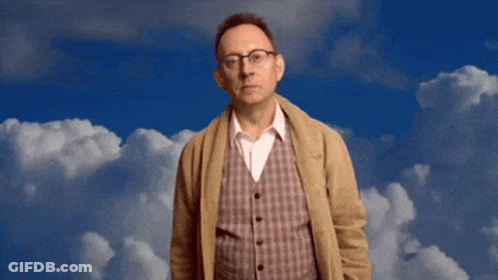 A Man Wearing Glasses And A Plaid Vest Stands In Front Of A Blue Sky With White Clouds GIF