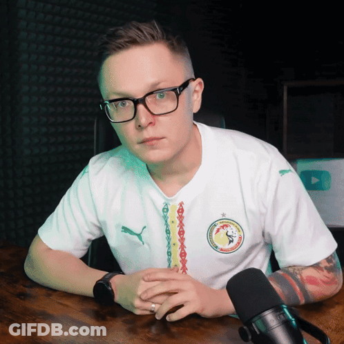A Man Wearing Glasses And A Puma Shirt Sits At A Table With A Microphone In Front Of Him GIF