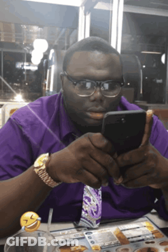 A Man Wearing Glasses And A Purple Shirt Is Looking At His Phone GIF