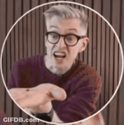 A Man Wearing Glasses And A Purple Shirt Is Pointing At The Camera . GIF