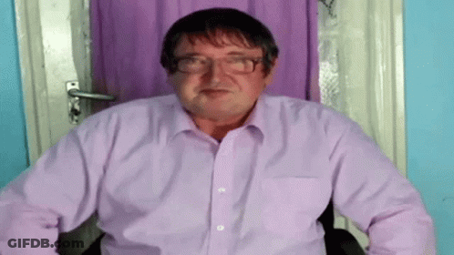 A Man Wearing Glasses And A Purple Shirt Is Sitting In A Chair . GIF