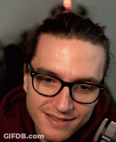 A Man Wearing Glasses And A Red Hoodie Is Smiling In Front Of A Rode Microphone GIF