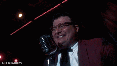 A Man Wearing Glasses And A Red Jacket Is Smiling In Front Of A Microphone GIF