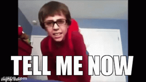 A Man Wearing Glasses And A Red Sweater Is Standing In Front Of A Sign That Says Tell Me Now GIF