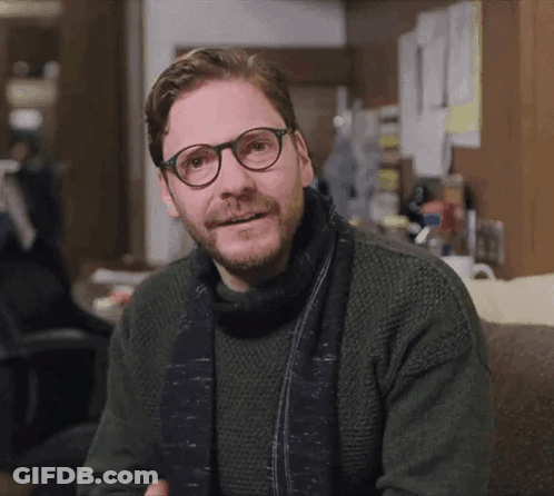 A Man Wearing Glasses And A Scarf Is Sitting On A Couch GIF