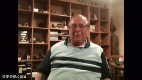 A Man Wearing Glasses And A Striped Shirt Is Sitting In Front Of A Bookshelf . GIF