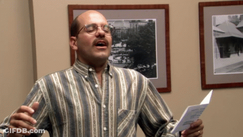 A Man Wearing Glasses And A Striped Shirt Sings Into A Microphone GIF
