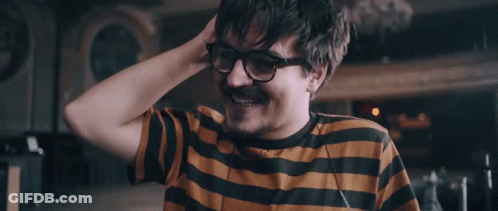 A Man Wearing Glasses And A Striped Shirt Smiles With His Hand In His Hair GIF
