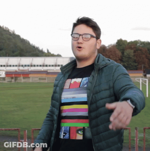 A Man Wearing Glasses And A Striped Shirt Stands In Front Of A Soccer Field GIF
