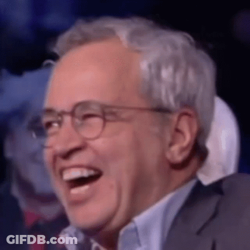 A Man Wearing Glasses And A Suit Is Laughing In A Crowd . GIF