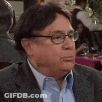 A Man Wearing Glasses And A Suit Is Sitting At A Table With Other People . GIF