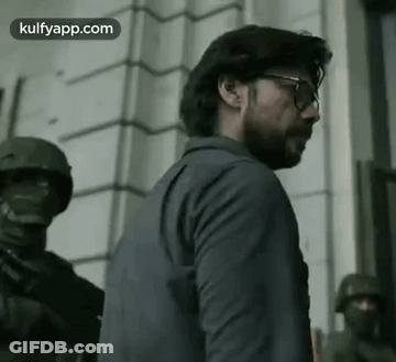 A Man Wearing Glasses And A Suit Is Standing In Front Of A Building With Soldiers . GIF