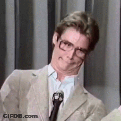 A Man Wearing Glasses And A Suit Is Standing In Front Of A Microphone Making A Funny Face . GIF