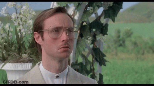 A Man Wearing Glasses And A Suit Stands In Front Of A Flower Pot GIF